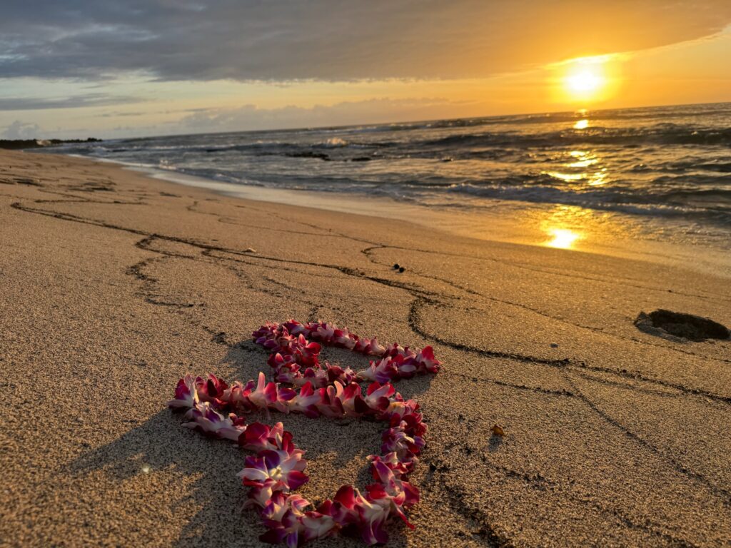 Golden sand beach, two purple and white orchid lei in the shape of hearts on the sand, gentle waves lapping on the sand, setting sun, on a blue, yellow, and orange sky.