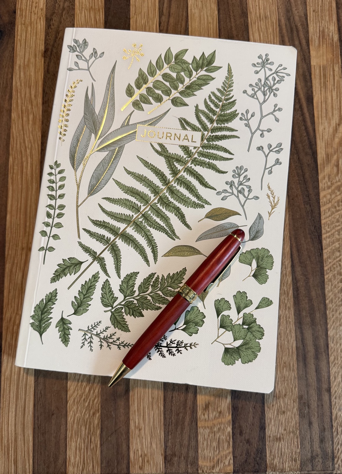 “Botanical journal with gold‑foil leaf illustrations and a red pen resting on top, placed on a wooden surface.